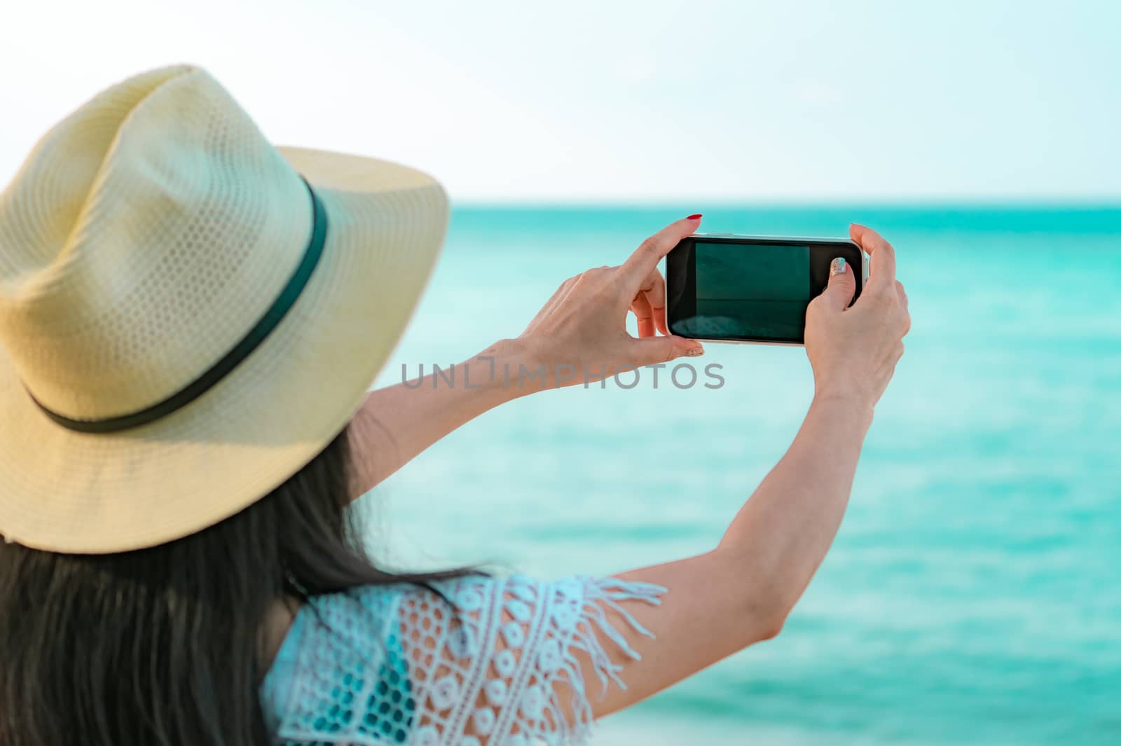 Young Asian woman wear hat use smartphone taking photo at tropical beach. Summer vacation at tropical paradise beach. Happy hipster girl travel on holiday. Woman enjoy and relax life. Summer vibes. 
