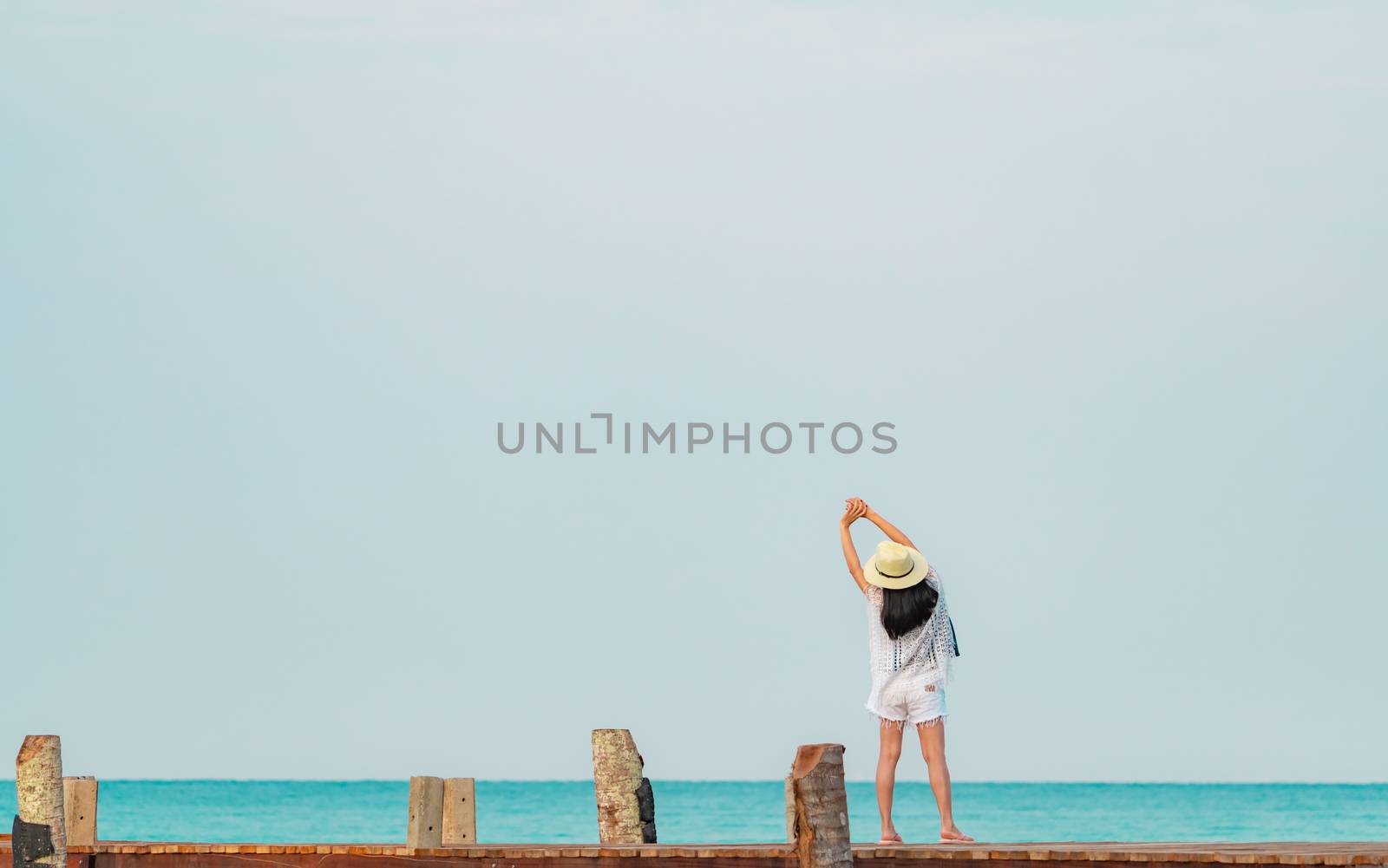 Back view of happy young Asian woman in casual style fashion and straw hat relax and enjoy holiday at tropical paradise beach. Girl standing at the wooden pier of resort in summer vacation fashion.