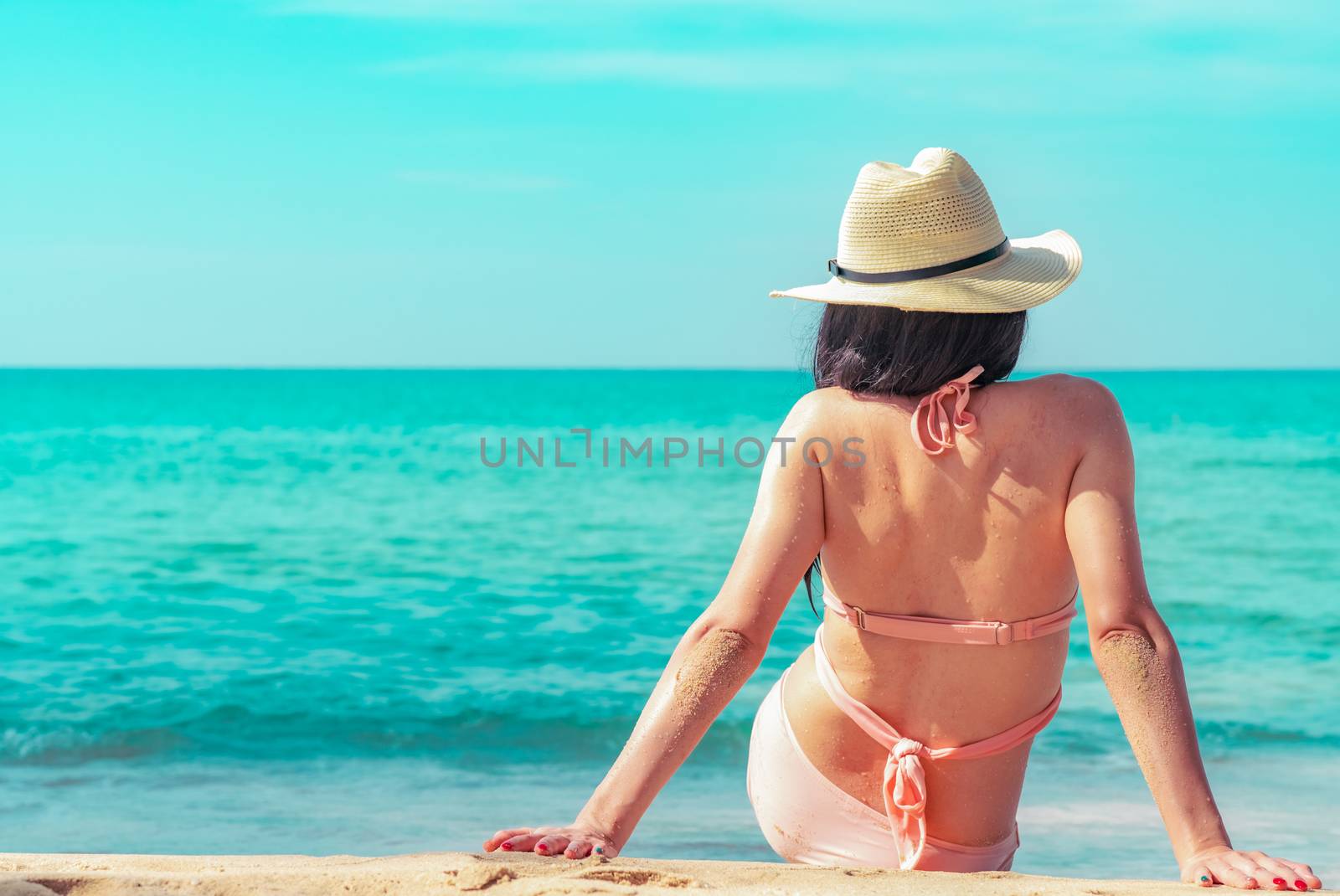 Back view of happy young Asian woman in pink swimsuit and straw hat relax and enjoy holiday at tropical paradise beach at sunset. Girl in summer vacation fashion. Beauty sexy model. Summer vibes.