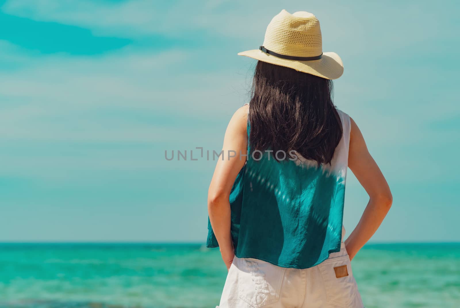 Happy young woman in casual style fashion and straw hat standing at sea beach. Relaxing and enjoy holiday at tropical paradise beach with emerald green water. Girl in summer vacation. Summer vibes.