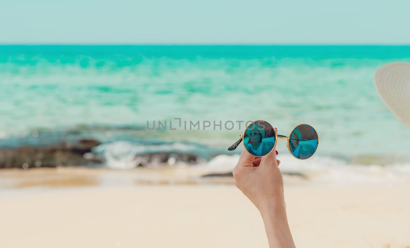 Woman hand holding sunglasses and sit on sand beach. Reflection of woman in sunglasses. Women wear straw hat relax at tropical beach on summer vacation. Sunny day on holiday. Travel alone on summer.