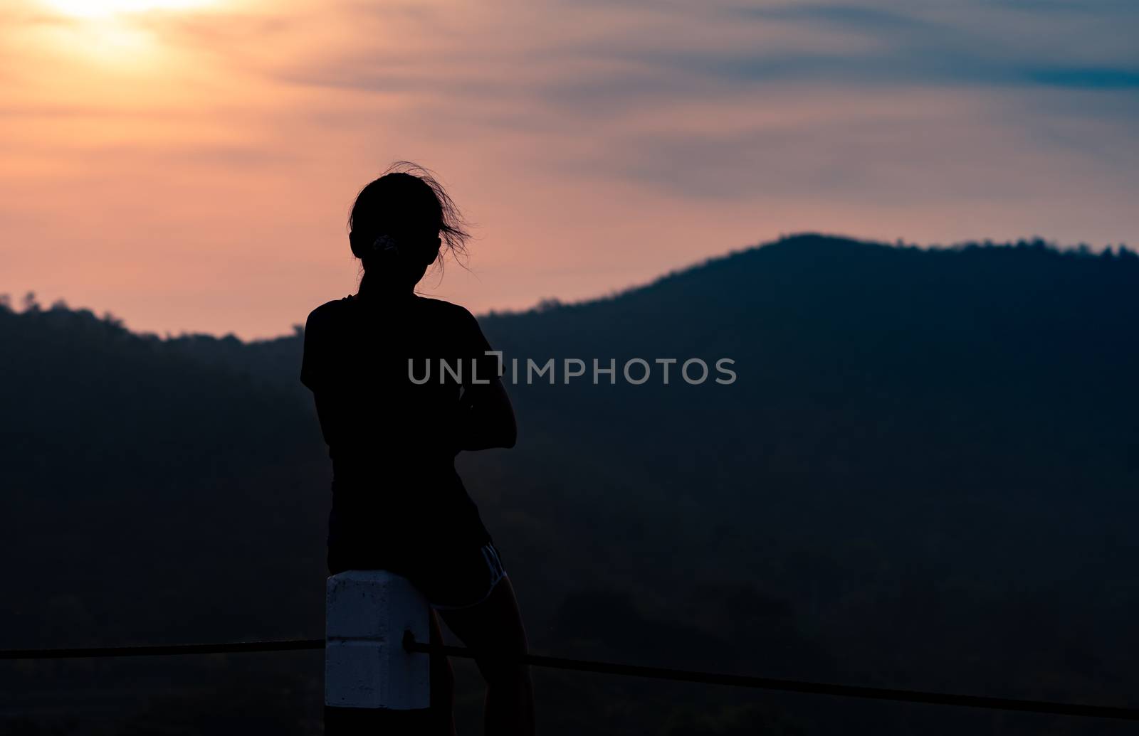Back view of a woman sit on fence post and watching sunrise above mountain in the morning. Adventure travel. Woman travel alone. People with peaceful mind by nature. Sitting to think about the past.