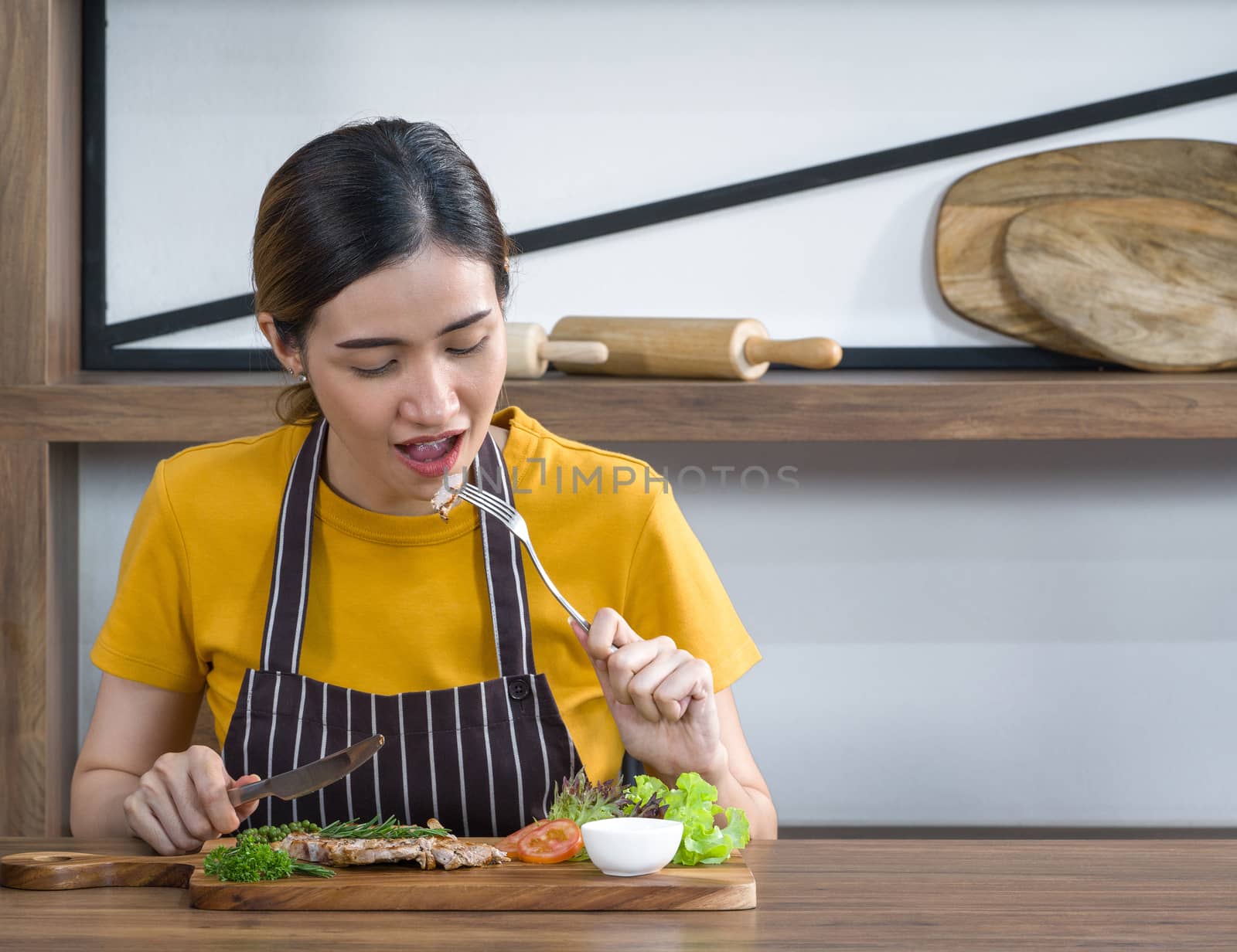 The housewife dressed in an apron enjoy eating the delicious steak. Morning atmosphere in a modern kitchen