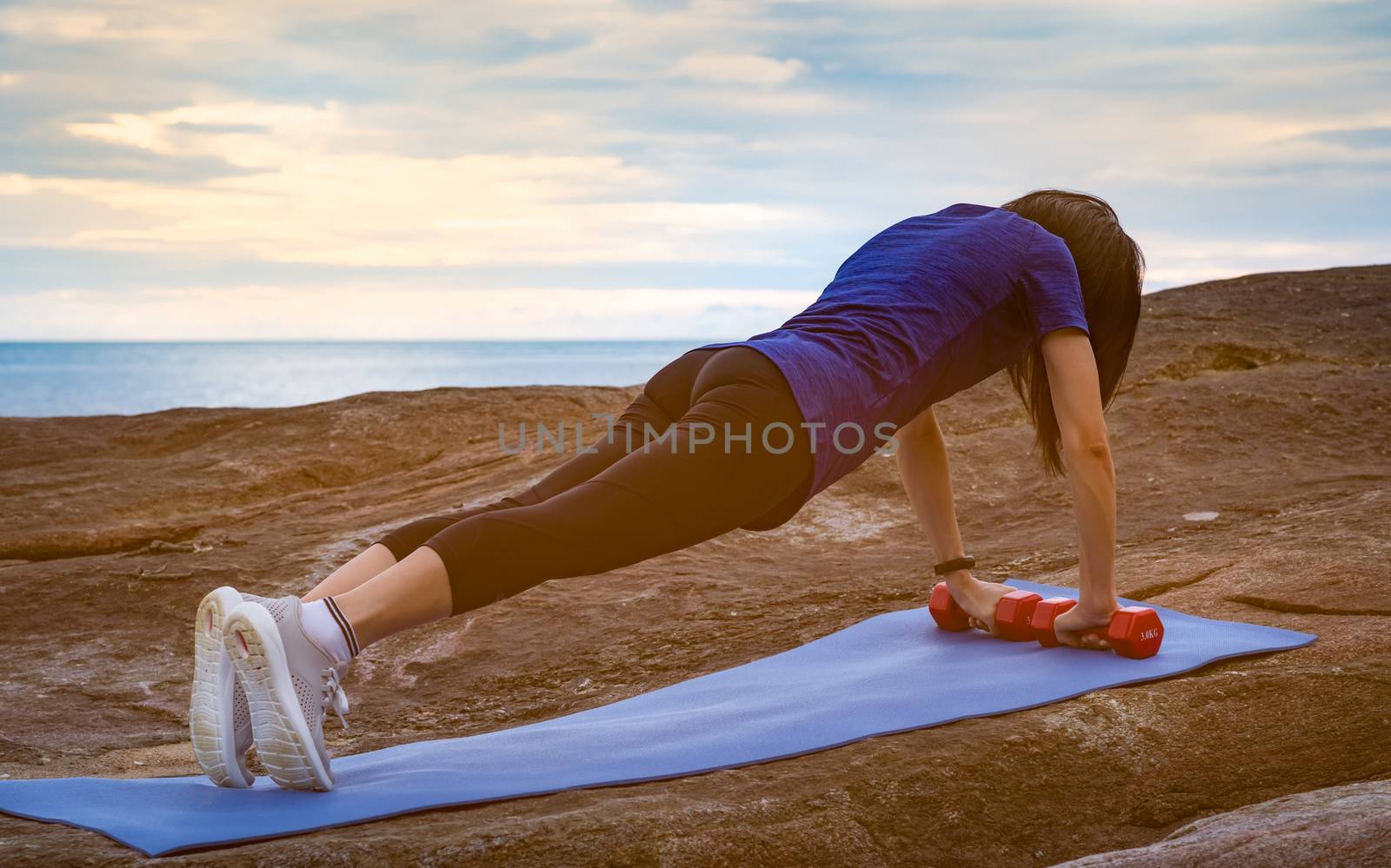 Asian woman workout in the morning at stone beach. Fit woman hand hold red dumbbell and push up on blue yoga mat for body strength. Weight training exercise for healthy lifestyle. Flat butt woman.