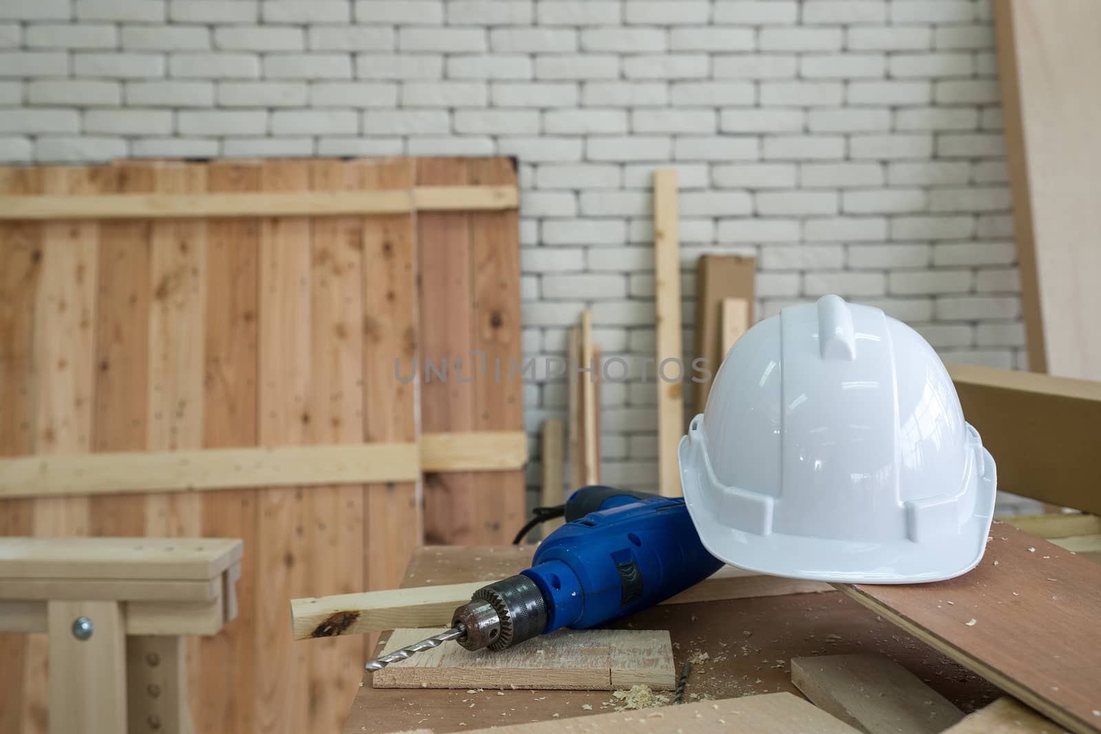 The white construction helmet and the blue electric drill were left aside during lunch time brake. A desk full of hand tools and wood piles. Morning work atmosphere in the workshop room.