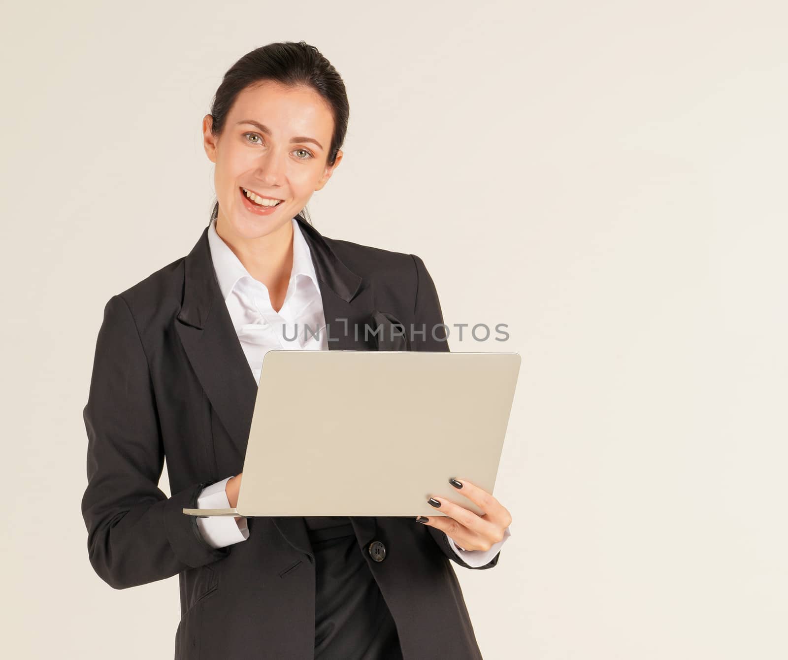 A business woman in a black suit with a smile and typing on a computer notebook. Portrait on beige background with studio light.