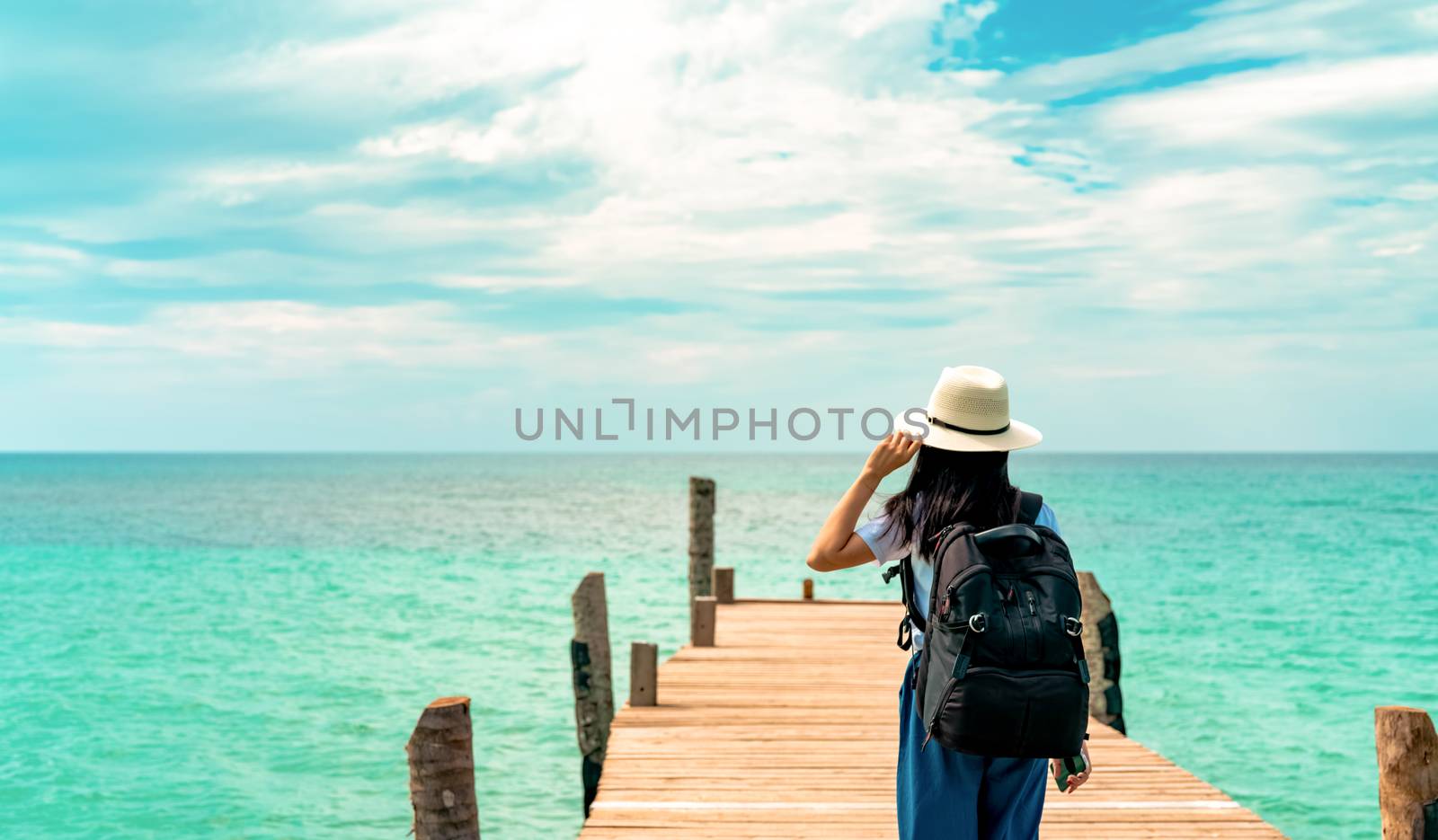 Happy young Asian woman in casual style fashion with straw hat and backpack. Relax and enjoy holiday at tropical paradise beach. Girl standing at the wooden pier of hotel in summer vacation fashion.