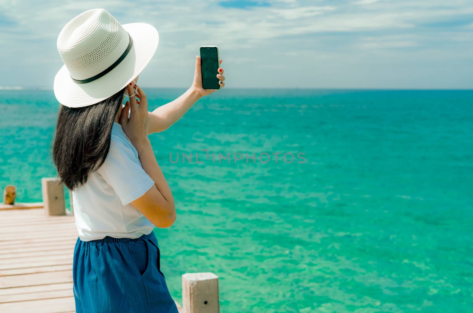 Young Asian woman wear hat in casual style use smartphone taking selfie at pier. Summer vacation at tropical paradise beach. Happy girl travel on holiday. Woman enjoy and relax life. Summer vibes. 