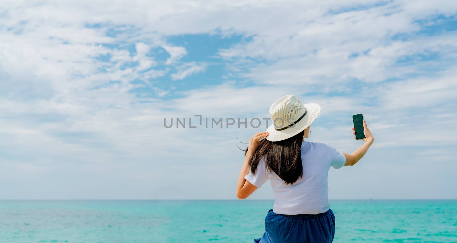 Young Asian woman wear straw hat use smartphone taking selfie at wooden pier. Summer vacation at tropical paradise beach. Happy girl travel on holiday. Woman enjoy and relax life. Summer vibes.