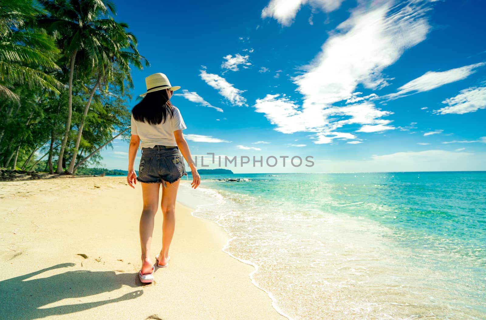 Back view of happy young woman in casual style fashion and straw hat wear sandals walking at sand beach. Relaxing and enjoy holiday at tropical paradise beach with blue sky. Girl in summer vacation. 