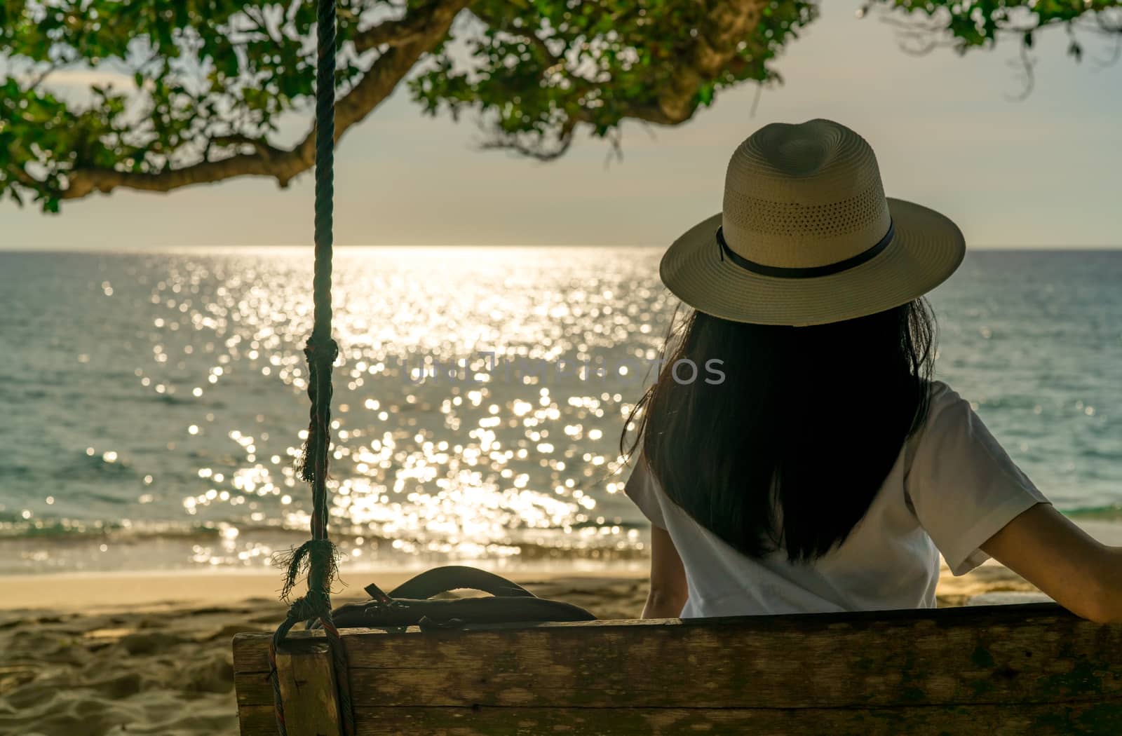 Young Asian woman sit and relax on swings at seaside on summer vacation. Summer vibes. Woman travel alone on holiday. Backpacker enjoy life at tropical paradise beach. Back view of woman watch sunset 
