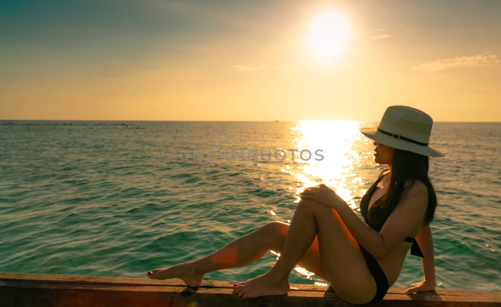 Sexy and relax woman wear black bikini with straw hat sit on wooden beam near sand beach at sunset. Girl enjoy holiday at tropical paradise beach on summer vacation. Holiday travel. Summer vibes.
