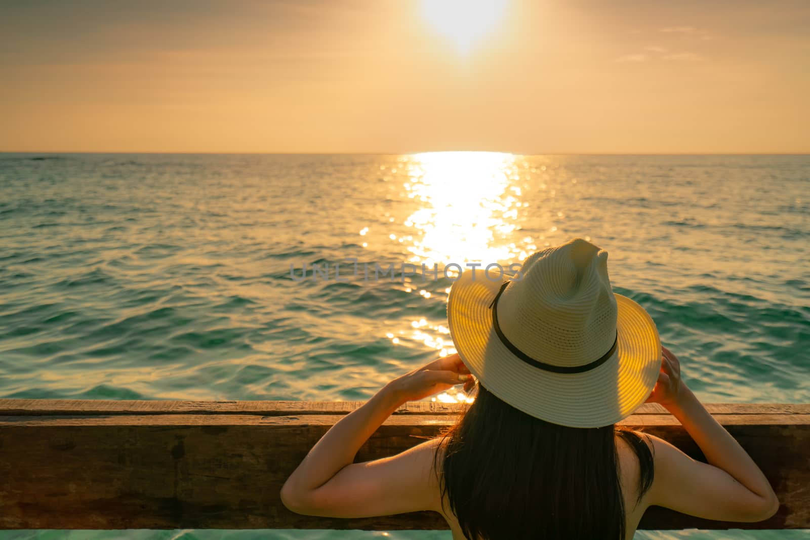 Back view of black long hair sexy Asian woman with straw hat relaxing and enjoying holiday at tropical paradise beach and watch the sunset sky. Summer vacation. Holiday travel alone. Summer vibes. 