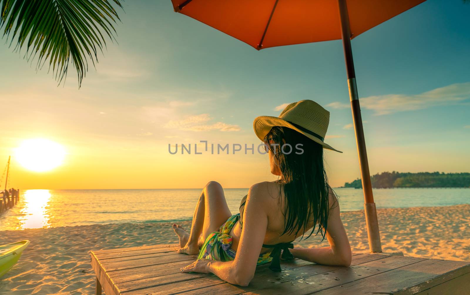 Sexy, enjoy and relax woman wear bikini lying and sunbathing on sunbed at sand beach at paradise tropical island under beach umbrella at sunset. Summer vacation. Holiday travel. Summer vibes.