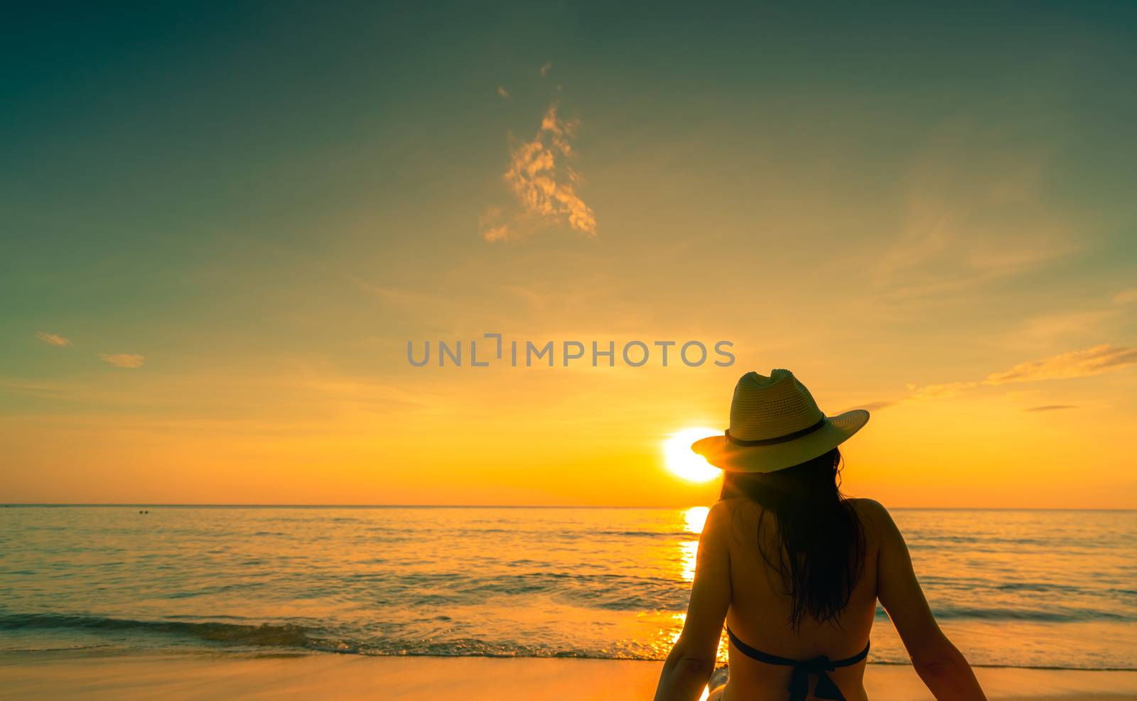 Back view of Asian woman wear bikini and straw hat sit on sand beach relax and enjoy holiday at tropical paradise beach. Woman watch beautiful sunset. Summer vacation. Travel alone. Summer vibes.