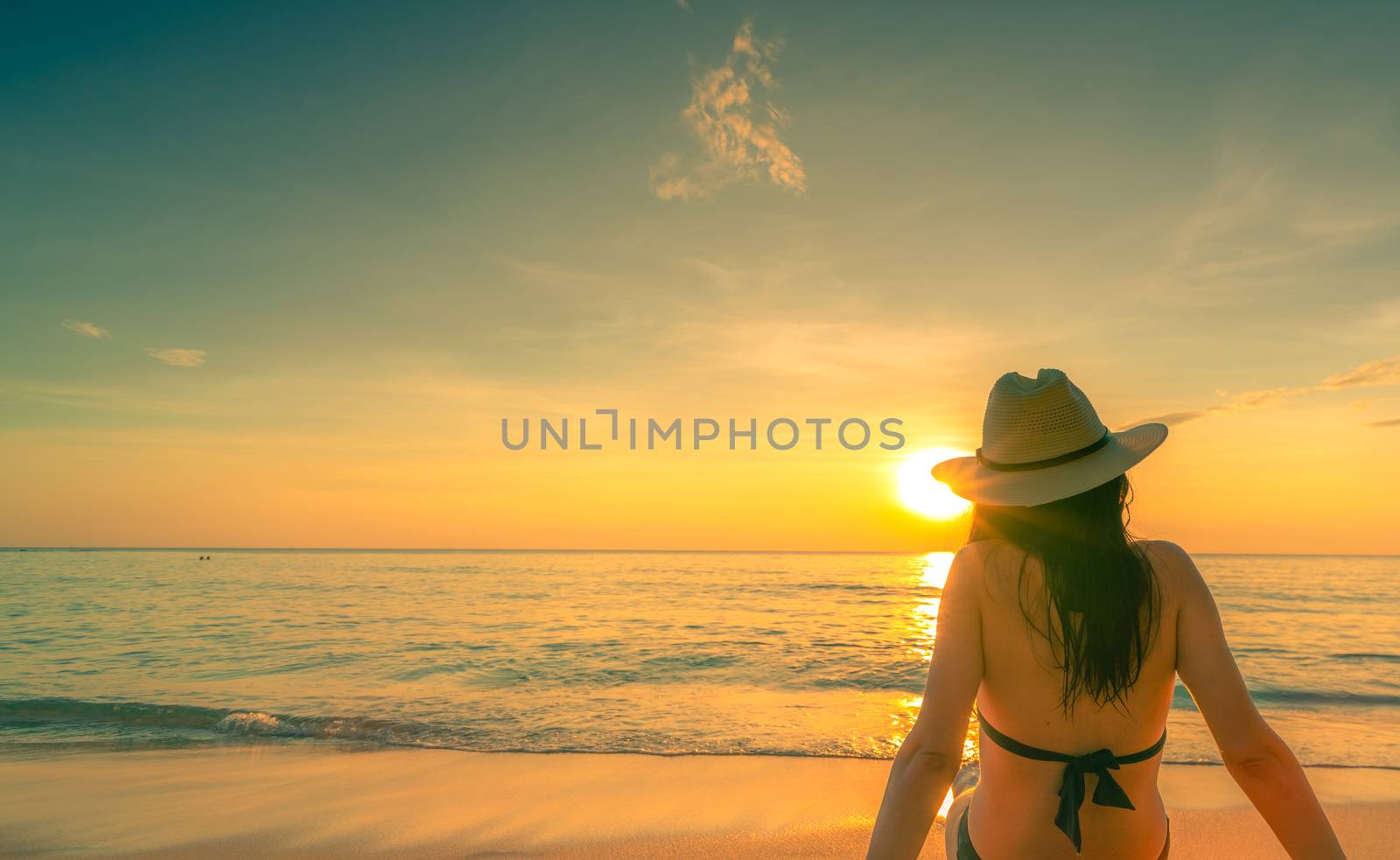 Back view of Asian woman wear bikini and straw hat sit on sand b by Fahroni