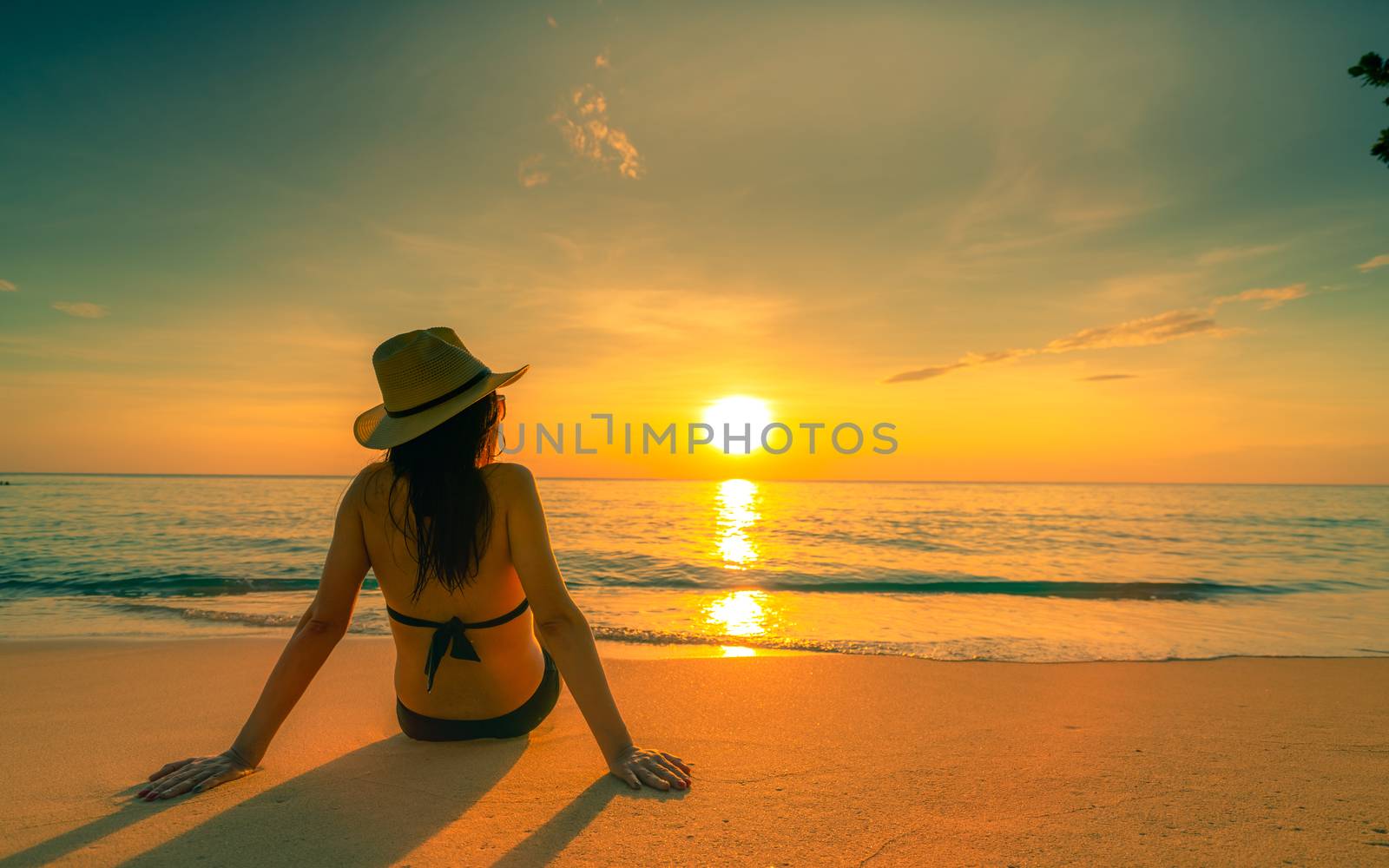 Back view of Asian woman wear bikini and straw hat sit on sand beach relax and enjoy holiday at tropical paradise beach. Woman watch beautiful sunset. Summer vacation. Travel alone. Summer vibes.