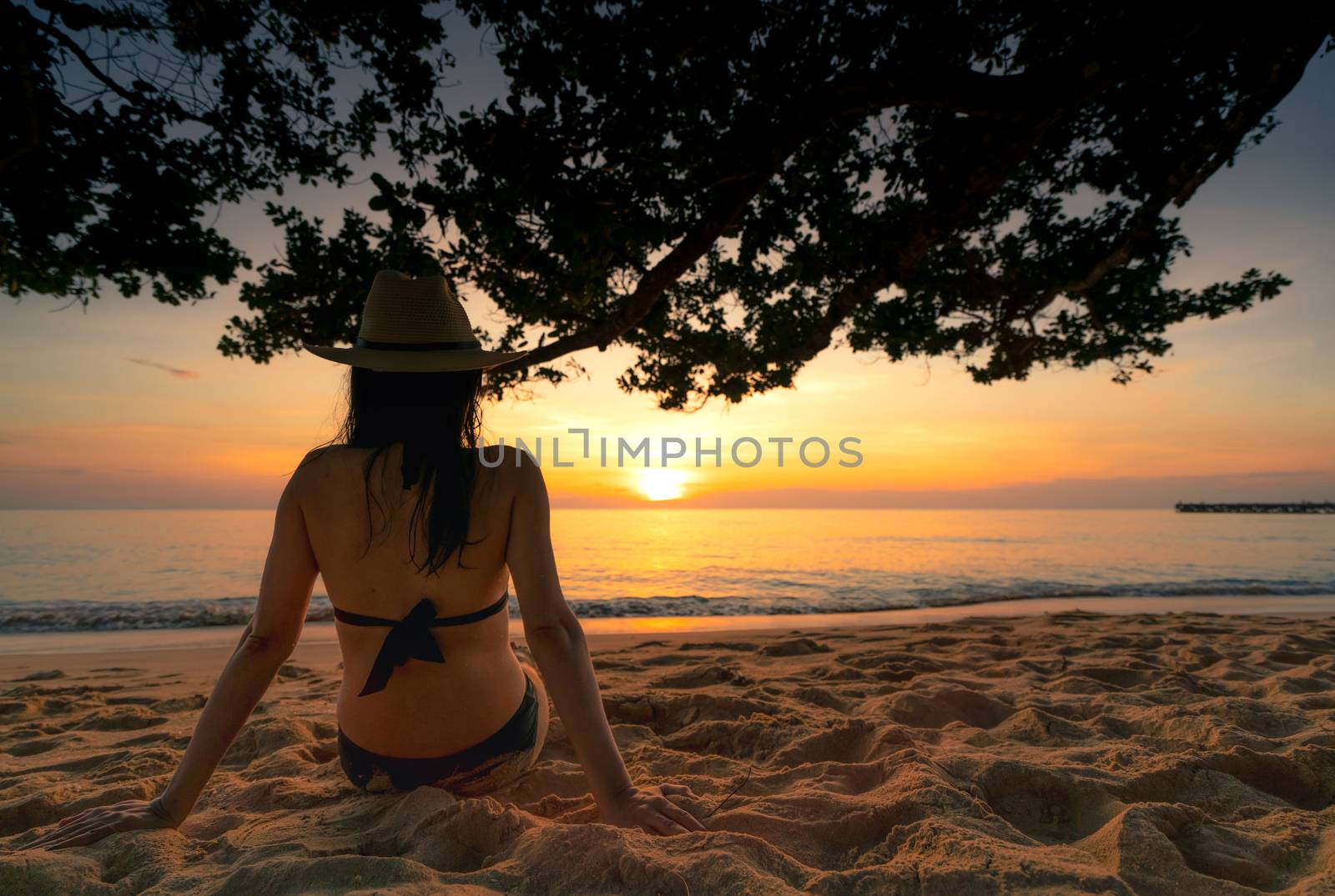 Back view of pregnant woman sit on sand and watching sunset at tropical beach. Woman wear swimsuit and straw hat relaxing at tropical paradise beach on summer vacation. Holiday travel alone.