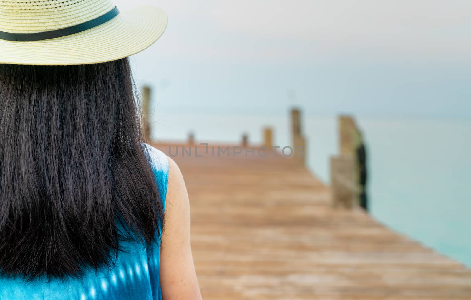Back view of happy young Asian woman in casual style fashion and straw hat relax and enjoy holiday at tropical paradise beach. Girl stand at the wooden pier of resort in summer vacation. Summer vibes.
