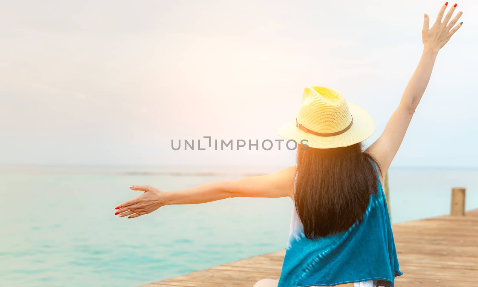 Back view of happy young Asian woman in casual style fashion and straw hat relax and enjoy holiday at tropical paradise beach. Girl stand at the wood pier of resort in summer vacation. Summer vibes.
