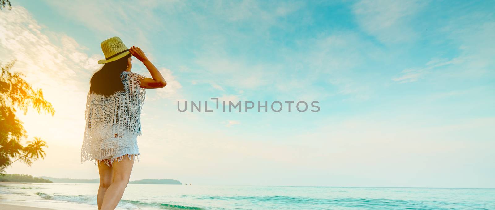 Happy young woman in casual style fashion and straw hat standing at sea beach. Relaxing and enjoy holiday at tropical paradise beach with emerald green sea water. Girl in summer vacation. Sunrise sky