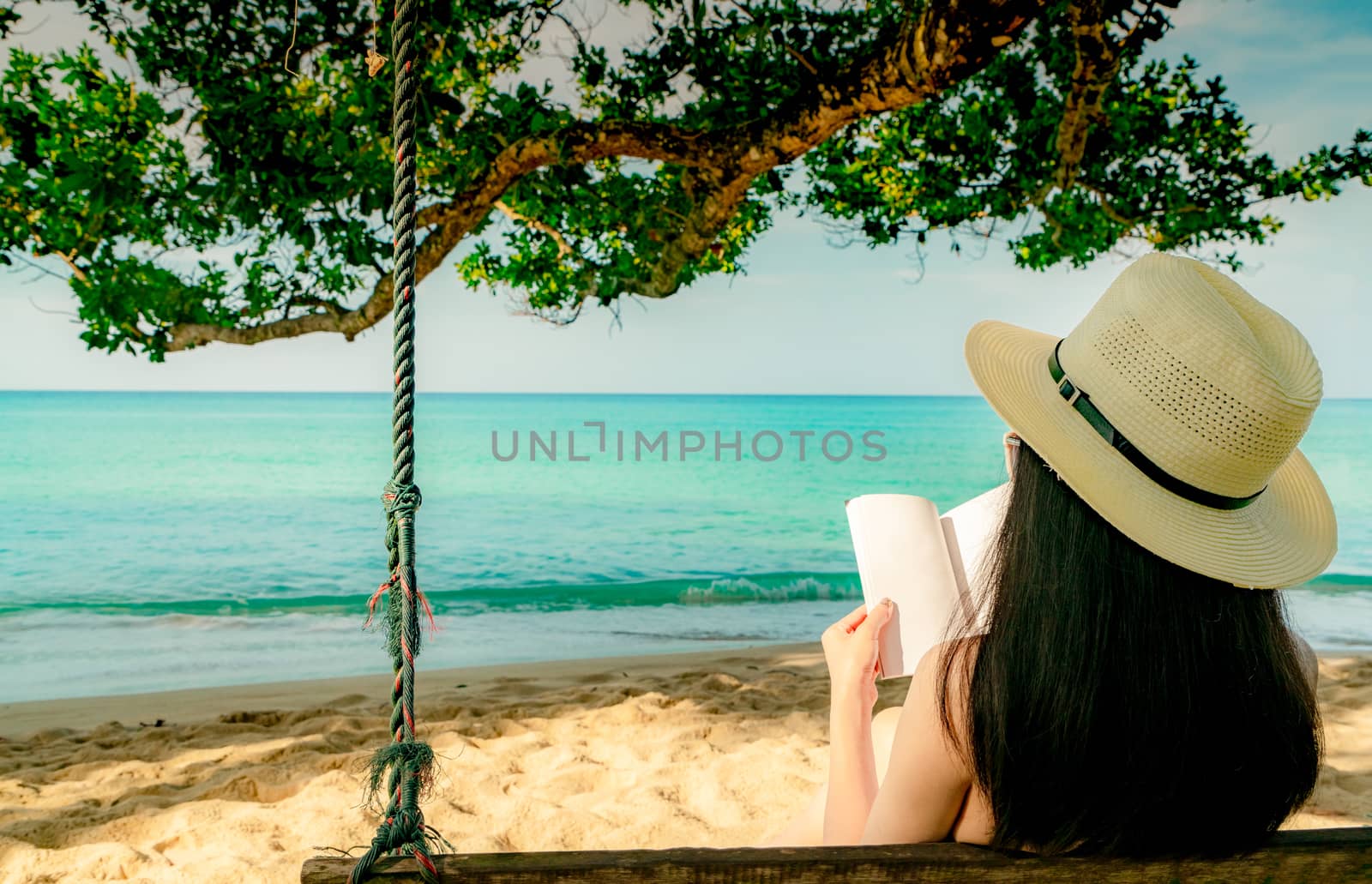 Women sit and reading a book on swings under the tree by the sea by Fahroni