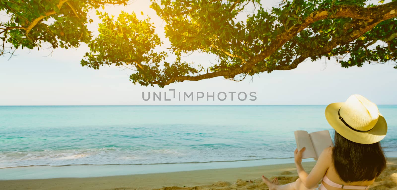 Women sit and reading a book under the tree at seaside. Back view of sexy Asian woman with straw hat relaxing and enjoying holiday at tropical paradise sand beach. Summer vacation. Summer vibes. 