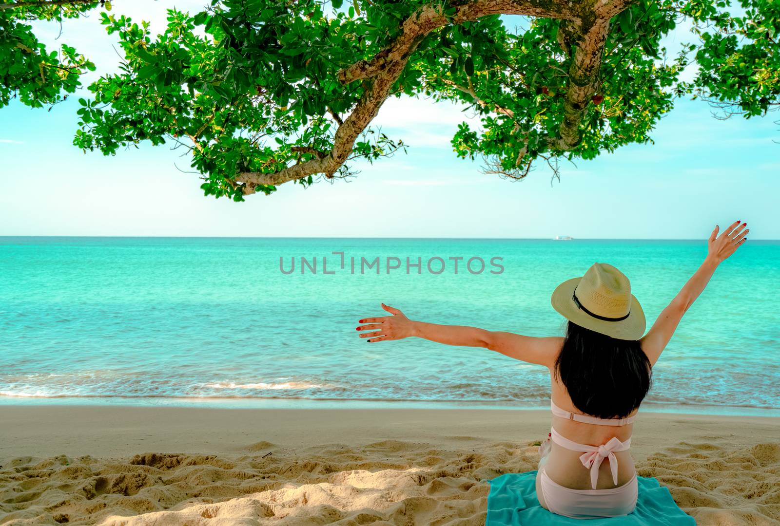 Back view of happy young Asian woman in pink swimsuit and straw hat relax and enjoy holiday at tropical sand beach under the tree. Girl in summer vacation fashion. Beauty sexy model. Summer vibes.