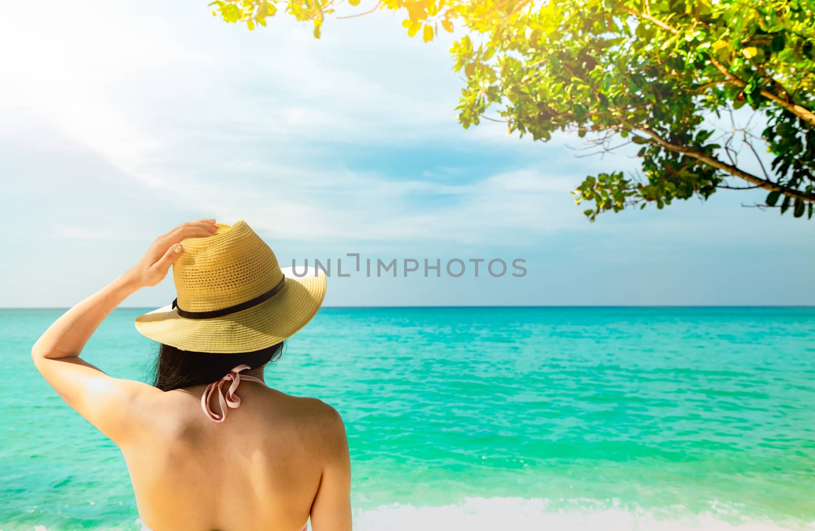 Back view of sexy and happy young Asian woman wear straw hat relaxing and enjoy holiday at tropical paradise beach. Girl standing at the beach and looking the sea in summer vacation. Summer vibes.