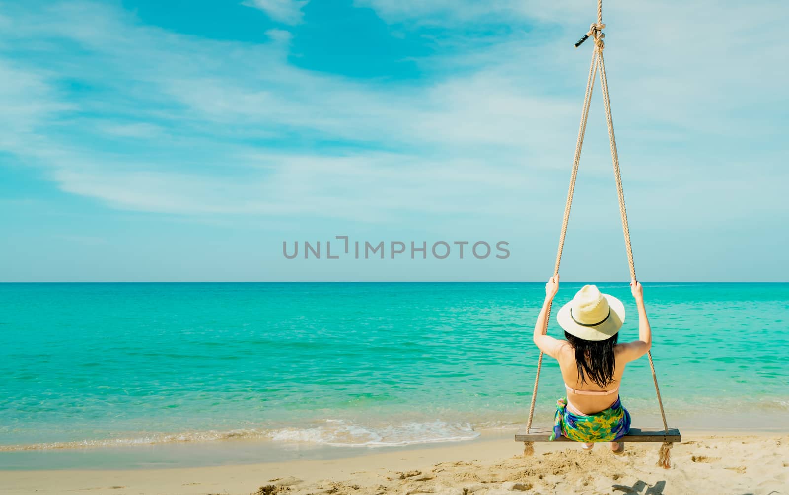 Asian woman wear swimwear and hat swing the swings at sand beach  and looking beautiful tropical paradise sea and sky on sunny day. Summer vacation. Summer vibes. Enjoying and relaxing girl on holiday