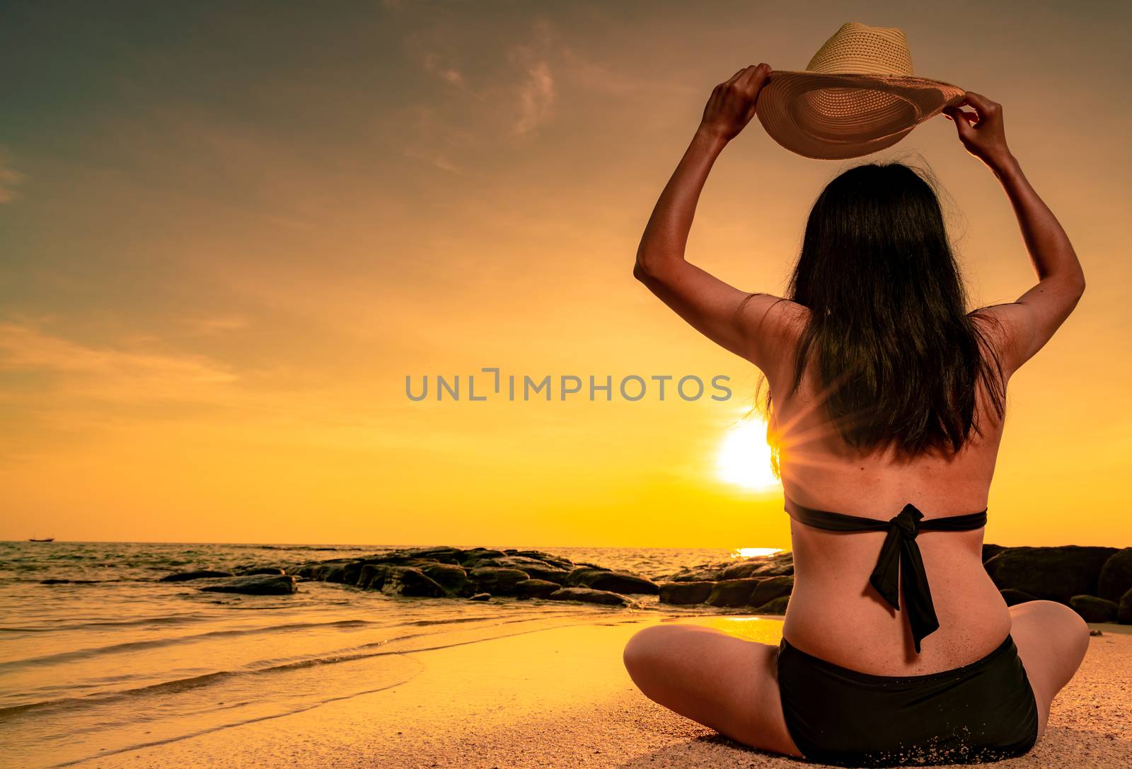 Back view of Asian plump woman wear bikini and hand holding straw hat sit on sand beach relax and enjoy holiday at tropical paradise beach. Woman watch beautiful sunset. Summer vacation. Summer vibes.