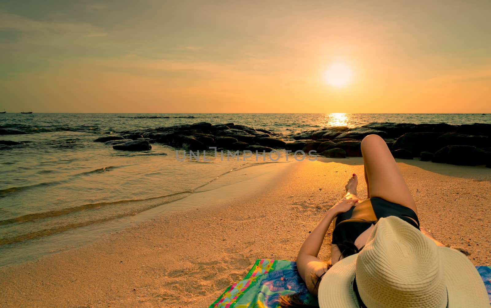 Woman lying down on sand beach at sunrise. Woman with straw hat sunbathing on tropical paradise beach with beautiful sunrise sky. Summer vacation. Girl wear black swimsuit sleep on sandy beach.