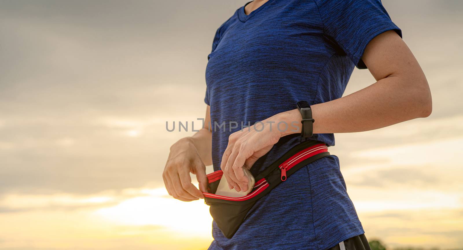 Young Asian woman keep smartphone in waist bag before running cardio exercise in the morning. Outdoor workout. Runner and smart band wearable device. Gadget for healthy lifestyle. Jogging fashion. 