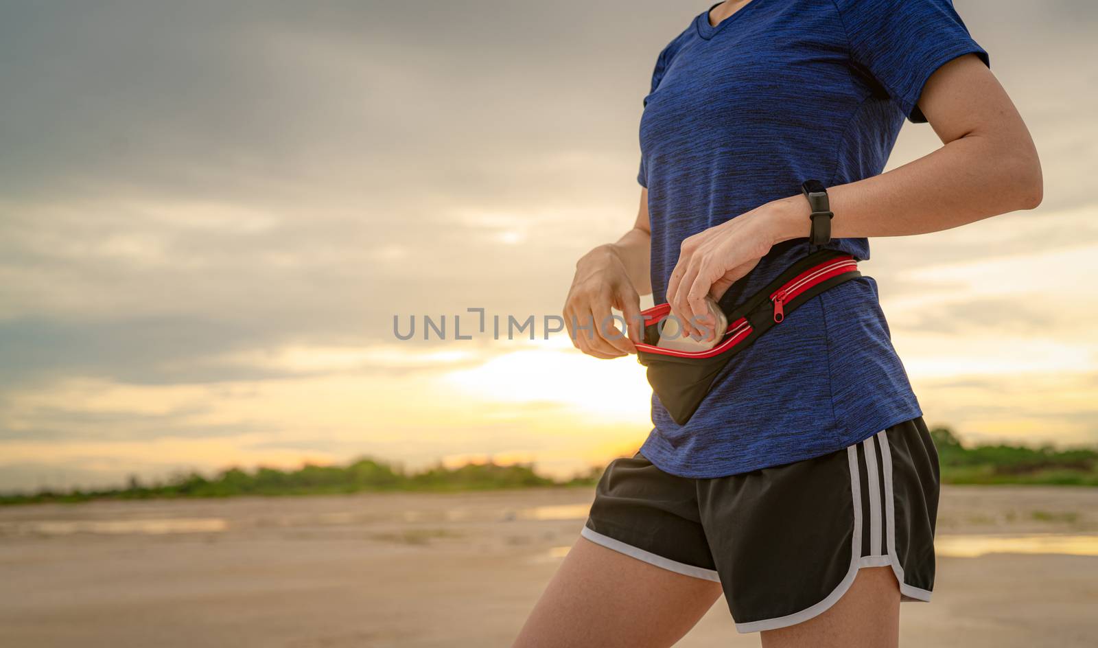 Young Asian woman keep smartphone in waist bag before running cardio exercise in the morning. Outdoor workout. Runner and smart band wearable device. Gadget for healthy lifestyle. Jogging fashion. 