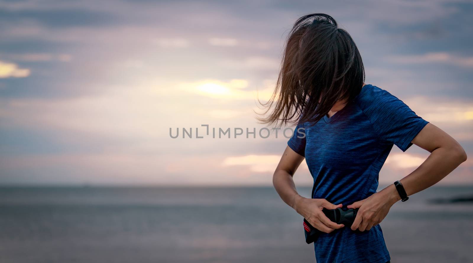 Asian woman wearing waist bag before running cardio exercise in the morning at sea beach with sunrise sky. Outdoor workout. Runner and smart band wearable device. Healthy lifestyle. Jogging fashion. 