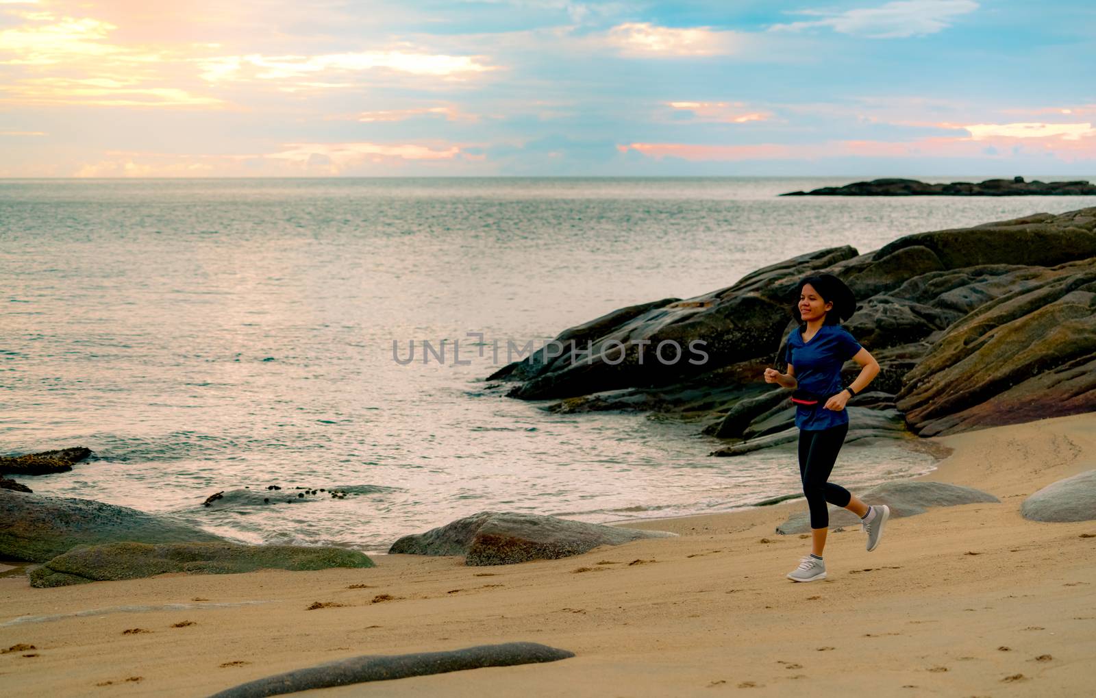 Asian woman running at tropical sea beach in the morning with beautiful sunrise sky. Fit healthy woman outdoor workout. Fit girl wear smart band and waist bag. Woman running for cardio exercise.