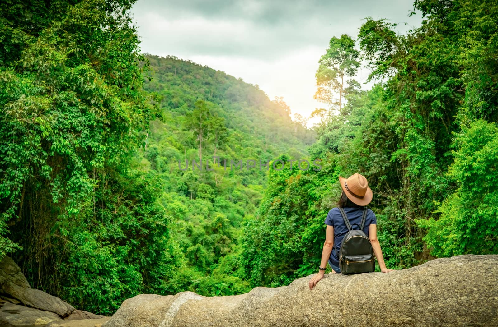 Happy woman travel alone in woods. Active woman with good mood sit on stone in green valley with dense tree in the forest. Happy vacation. Back view of happy woman wear backpack, smart band, and hat.