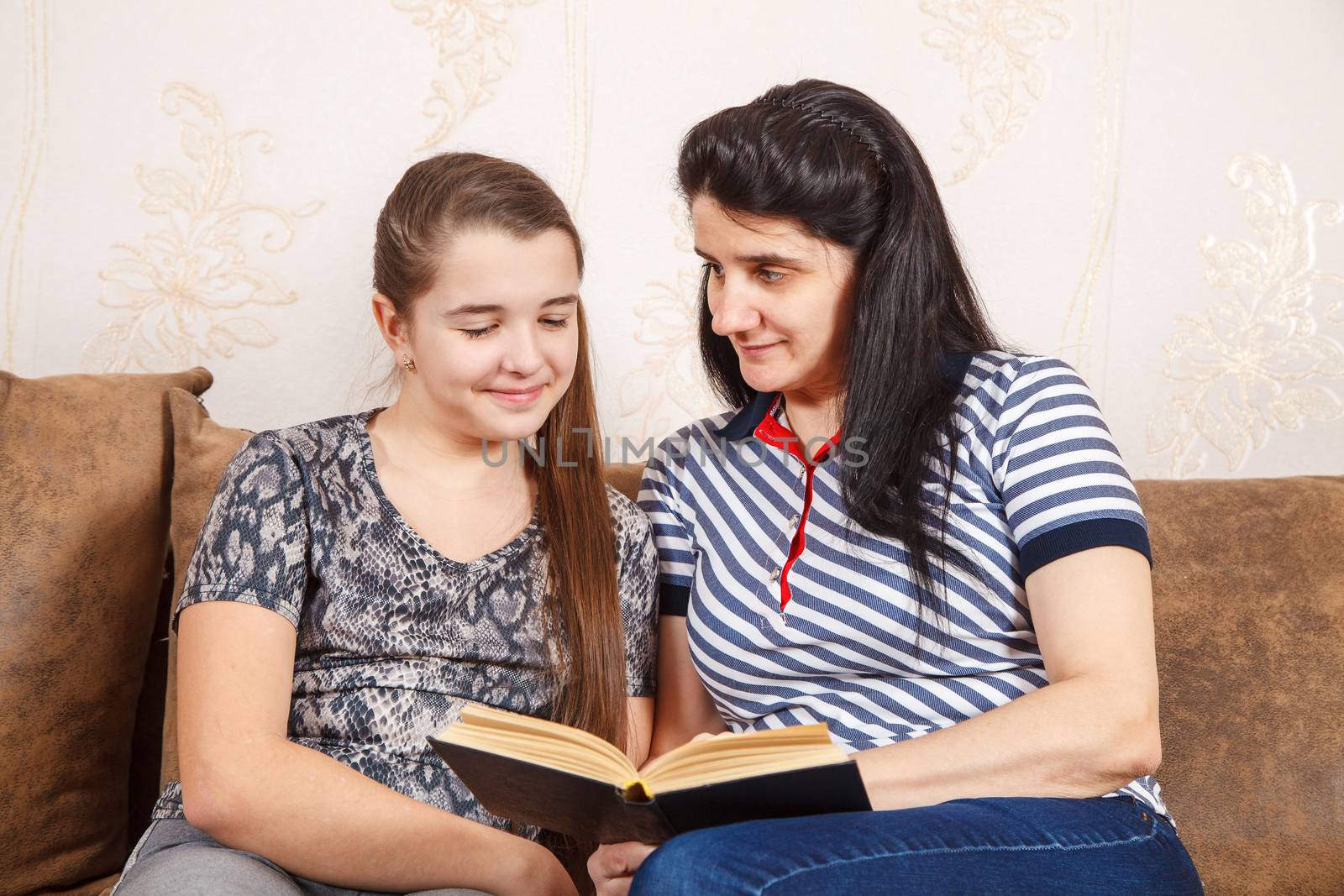 mom and daughter are reading a book while sitting on the sofa by raddnatt