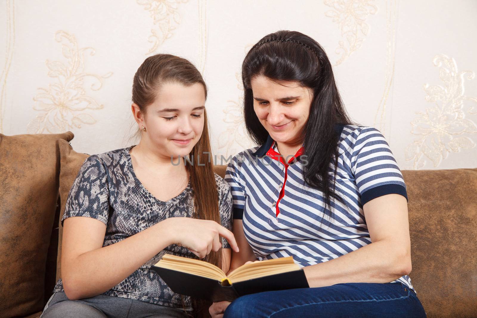 mom and daughter are reading a book while sitting on the sofa by raddnatt