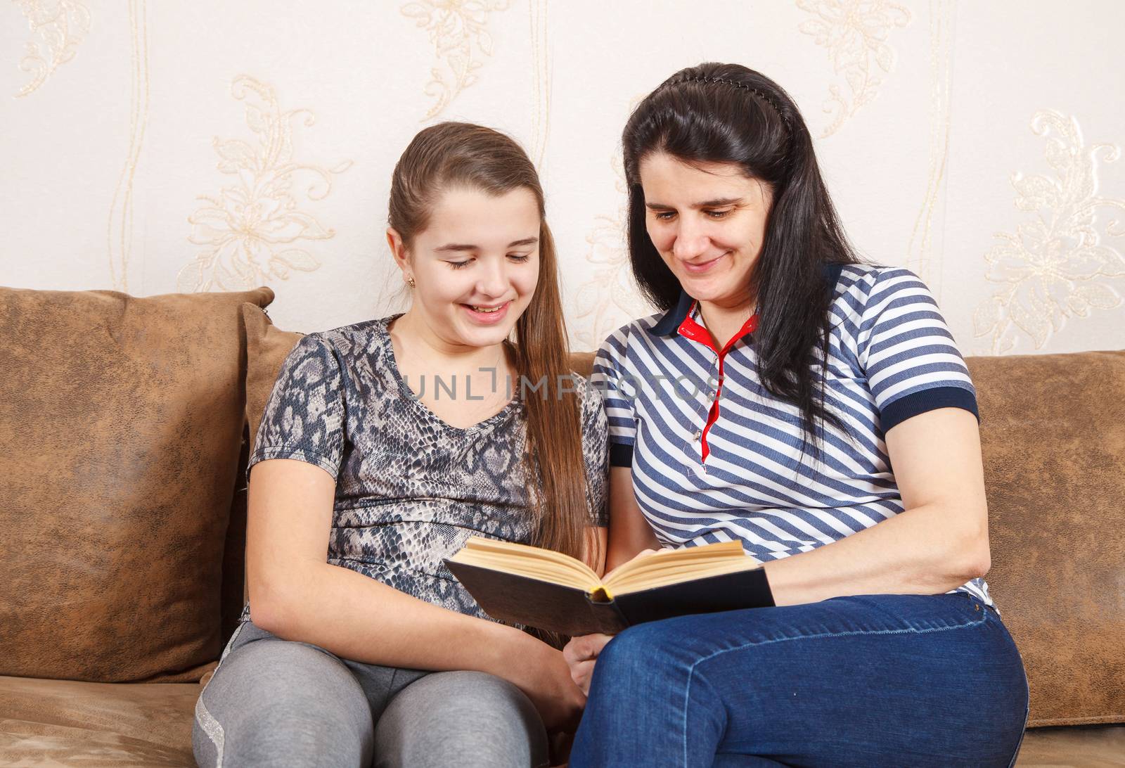 mom and daughter are reading a book while sitting on the sofa by raddnatt