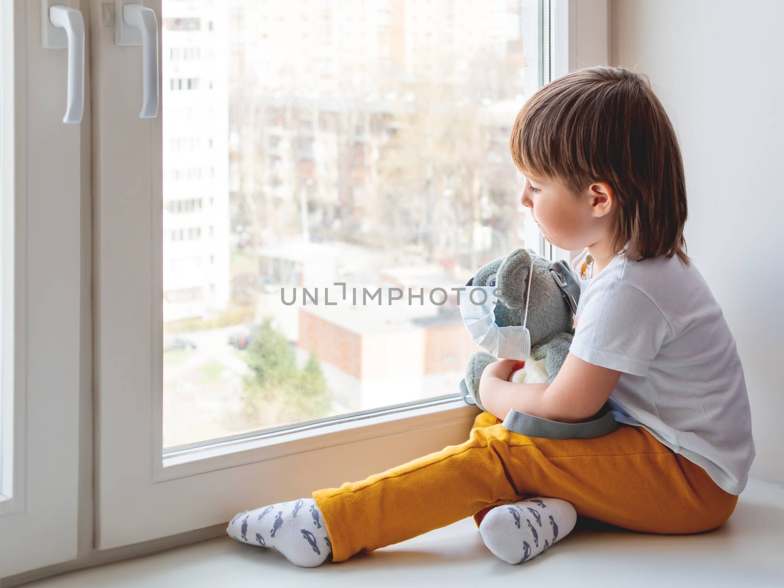 Toddler boy sits with teddy bear in medical mask. Kid with plush toy look through window outside. Koala bear and child on home quarantine because of coronavirus COVID-19.