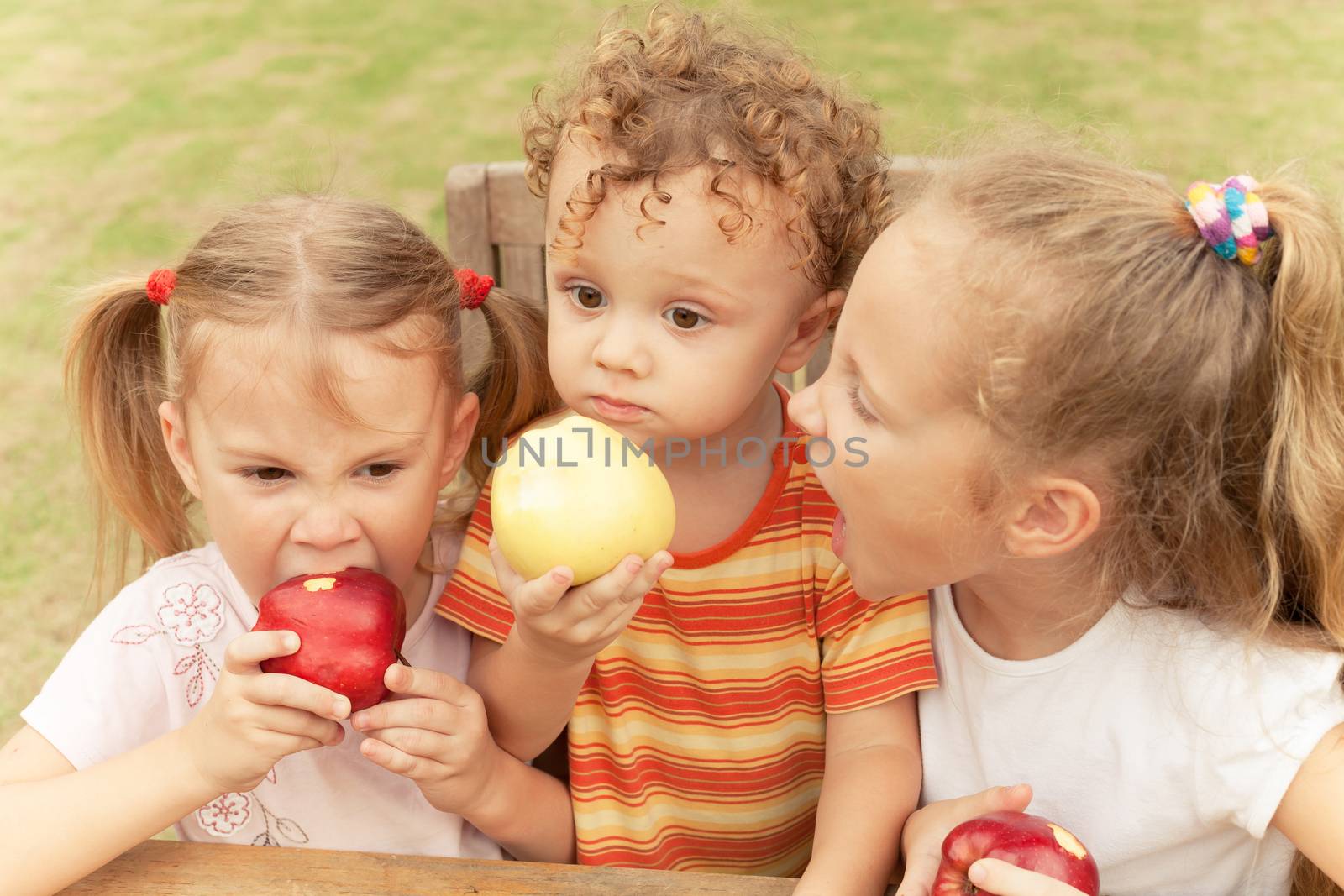 three happy children sitting at the table and eat apples by altanaka