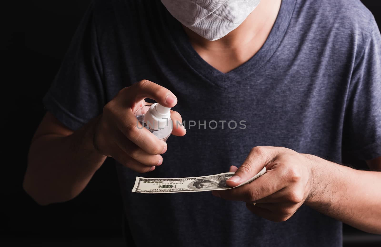 Closeup man wearing protective face mask applying sanitizer spray to 100 dollar money for cleaning, hygiene prevention COVID-19 virus or coronavirus protection concept, dark on black background
