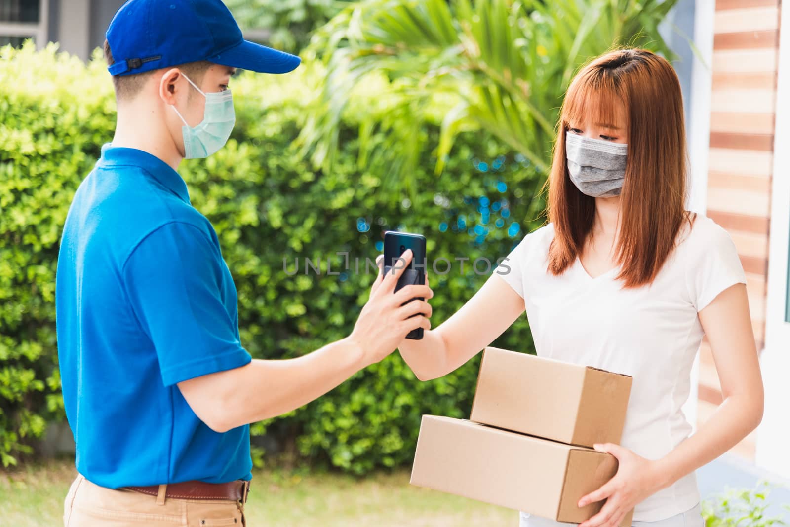 Asian delivery express courier young man giving parcel boxes to woman customer signature for receiving on mobile phone both protective face mask, under curfew quarantine pandemic coronavirus COVID-19