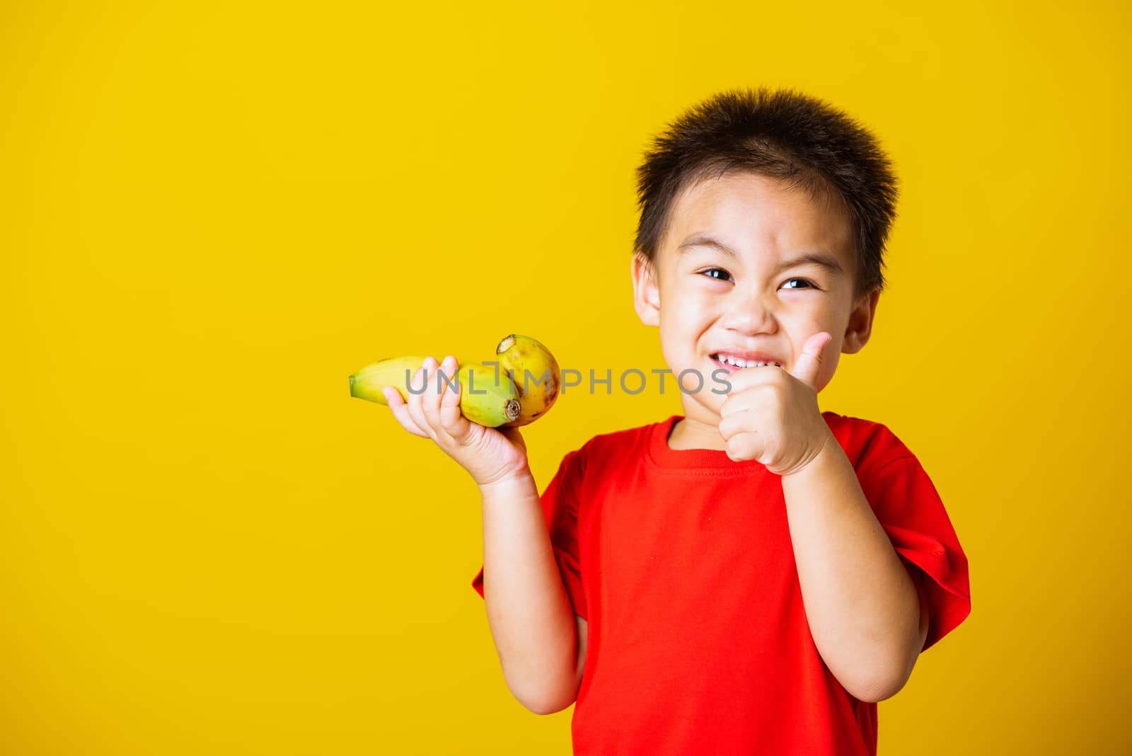 Happy portrait Asian child or kid cute little boy attractive smile wearing red t-shirt playing holds bananas and show finger thumb for good sign, studio shot isolated on yellow background
