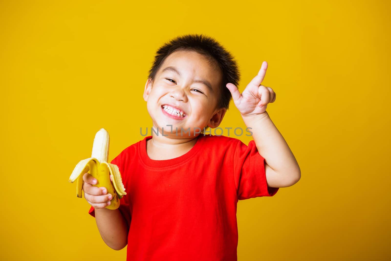 Happy portrait Asian child or kid cute little boy attractive smile wearing red t-shirt playing holds peeled banana for eating, studio shot isolated on yellow background