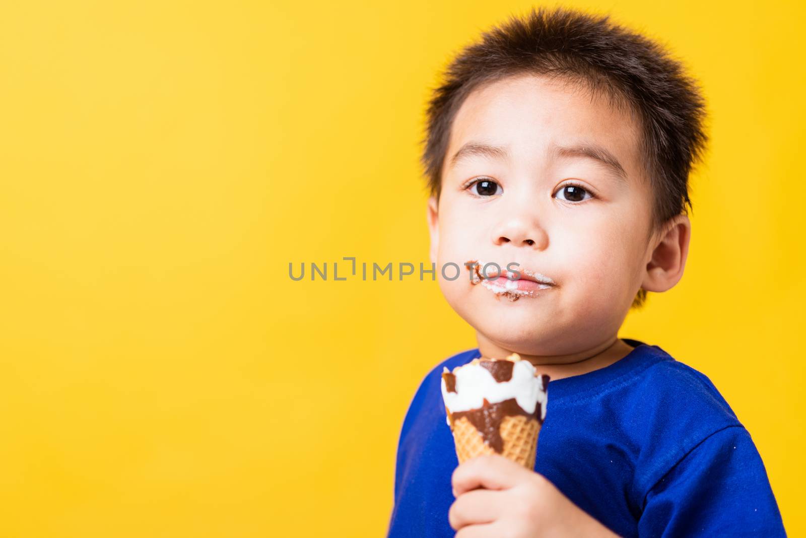 Happy portrait Asian child or kid cute little boy attractive laugh smile playing holds and eating sweet chocolate ice cream waffle cone, studio shot isolated on yellow background, summer concept