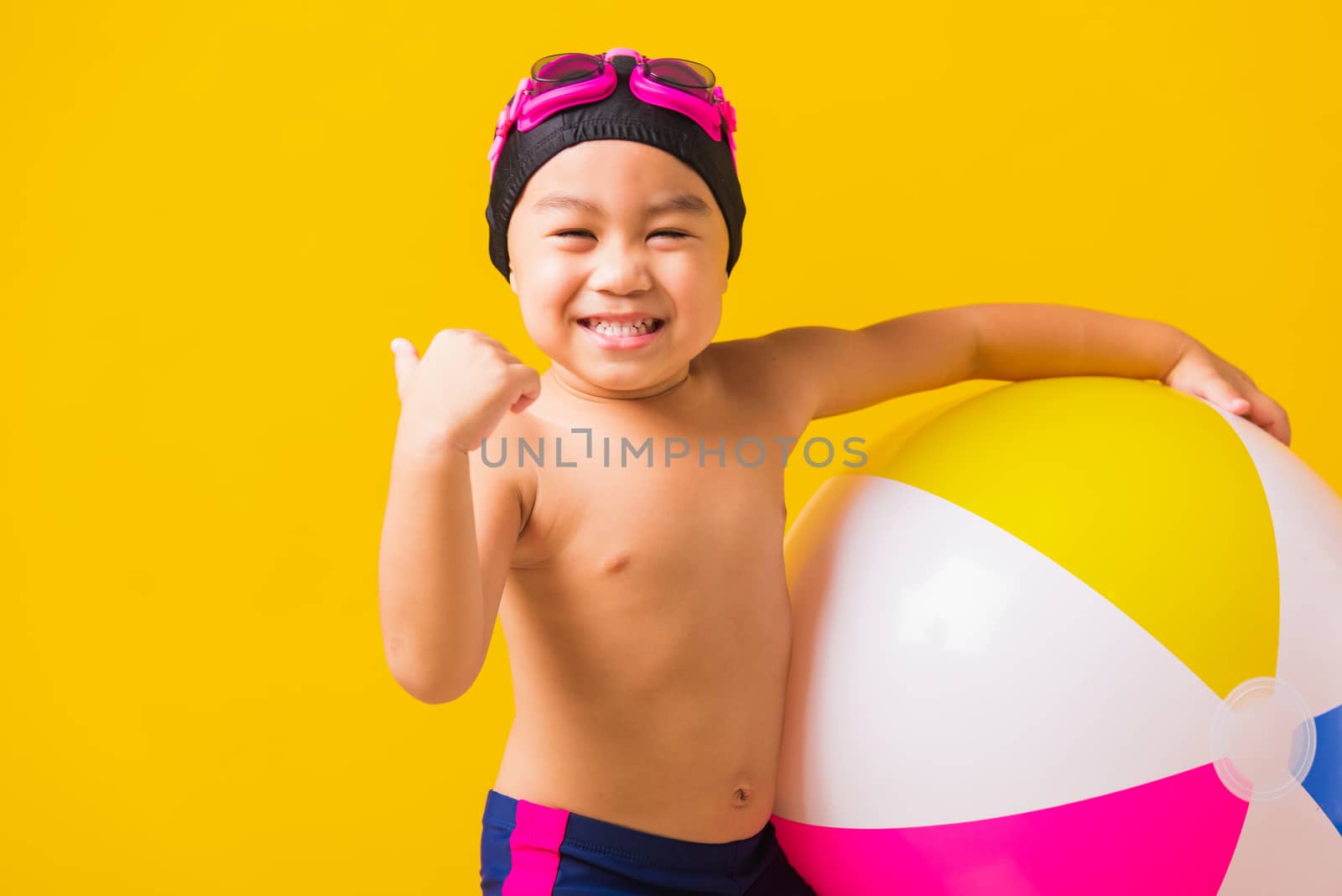 Summer vacation concept, Portrait Asian happy cute little child boy smiling in swimsuit hold beach ball and showing finger thumb up for good sign, studio shot isolated yellow background