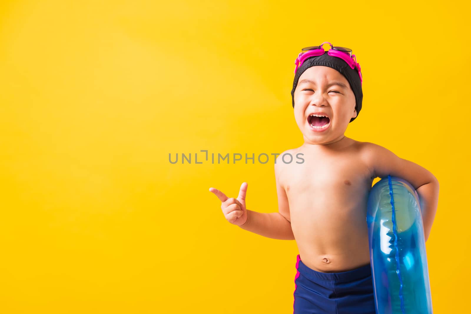 Summer vacation concept, Portrait Asian happy cute little child boy wear goggles and swimsuit hold blue inflatable ring, Kid hav fun point finger to side away, studio shot isolated yellow background