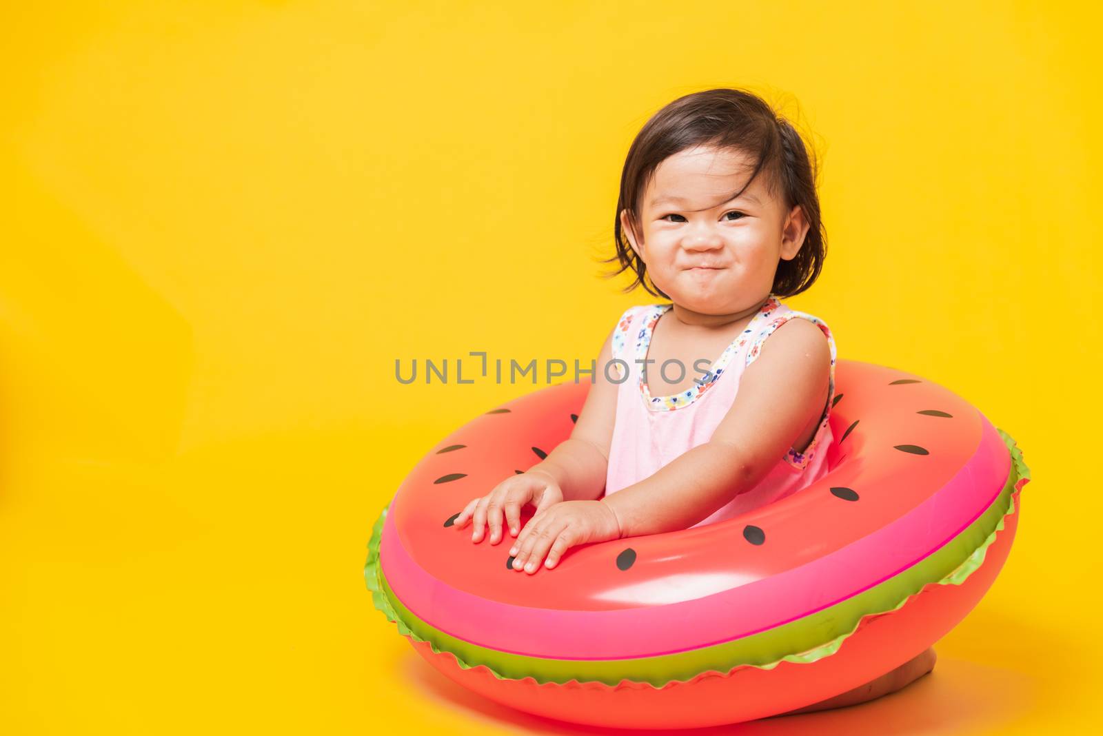 Summer vacation concept, Portrait Asian happy cute little baby girl wear swimsuit sitting in watermelon inflatable ring, Kid have fun sit in inflatable, studio shot isolated yellow background
